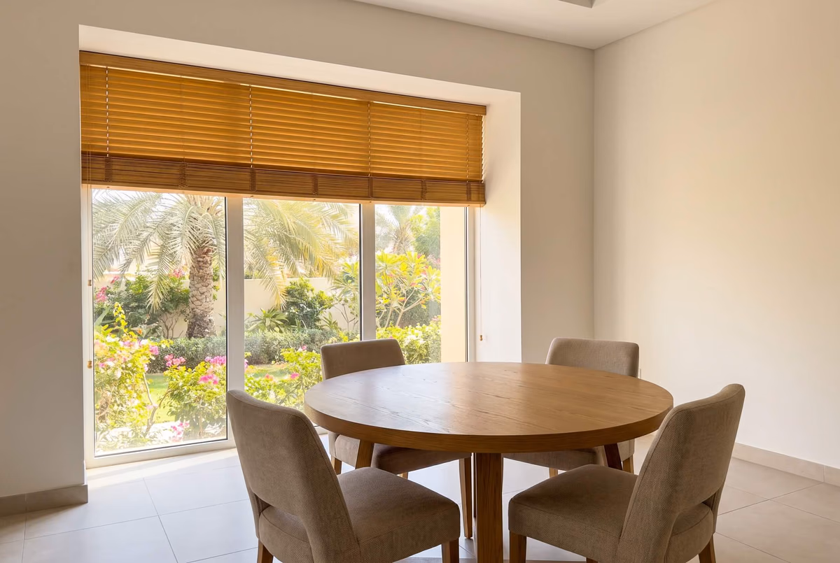 Villa dining room with a honey wooden blind raised to the top third of tall garden glass, the stacked slats covering the head of the window above a round oak table