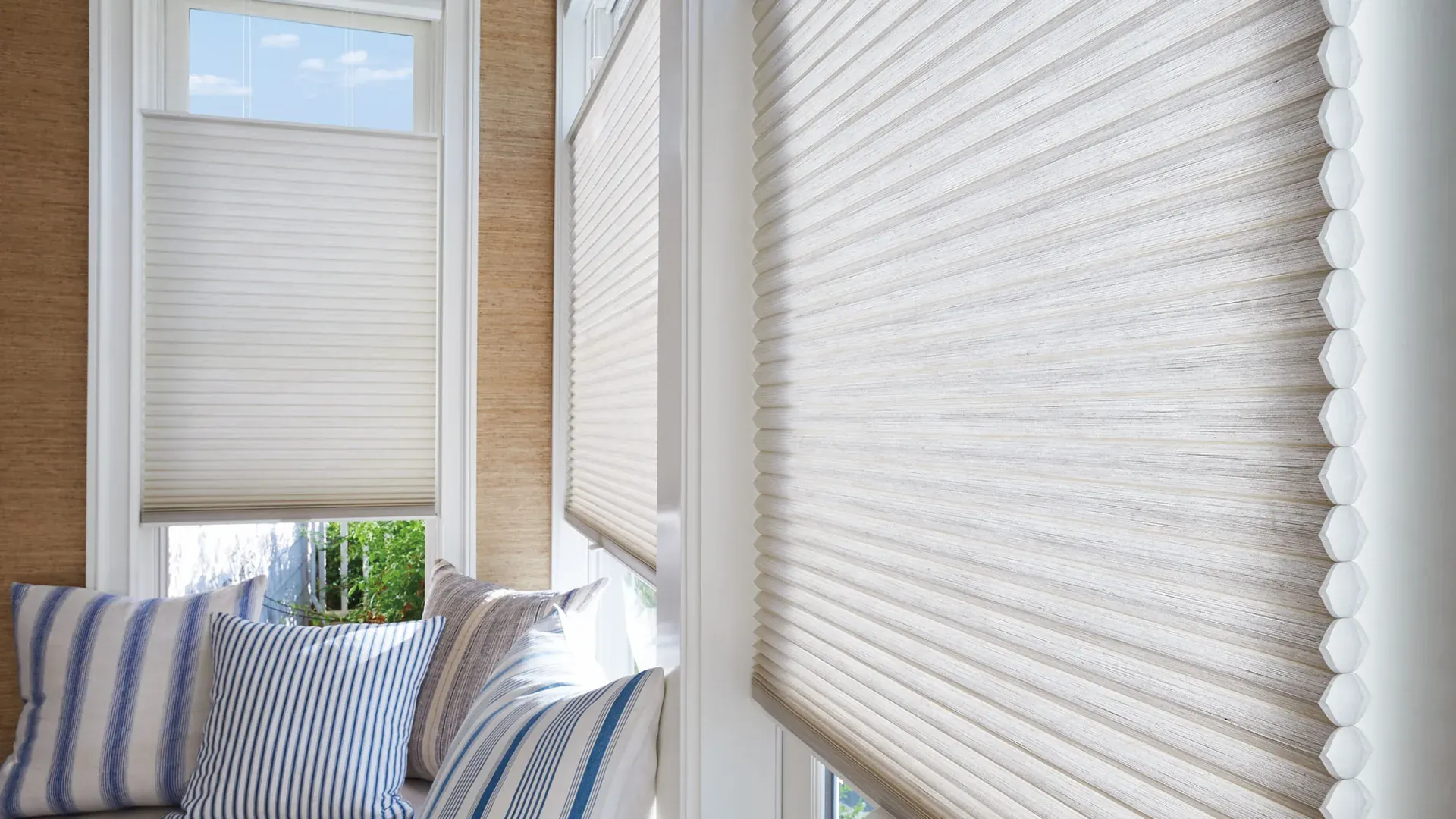 Cream honeycomb blinds part-lowered across the windows of a bay, the hexagonal cells visible edge-on at the nearest blind, above a window seat with blue striped cushions