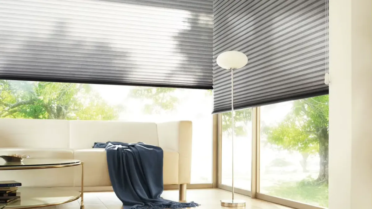 Grey honeycomb blinds lowered across two walls of corner windows in a room with a raised timber floor, daylight coming through the cloth rather than round it