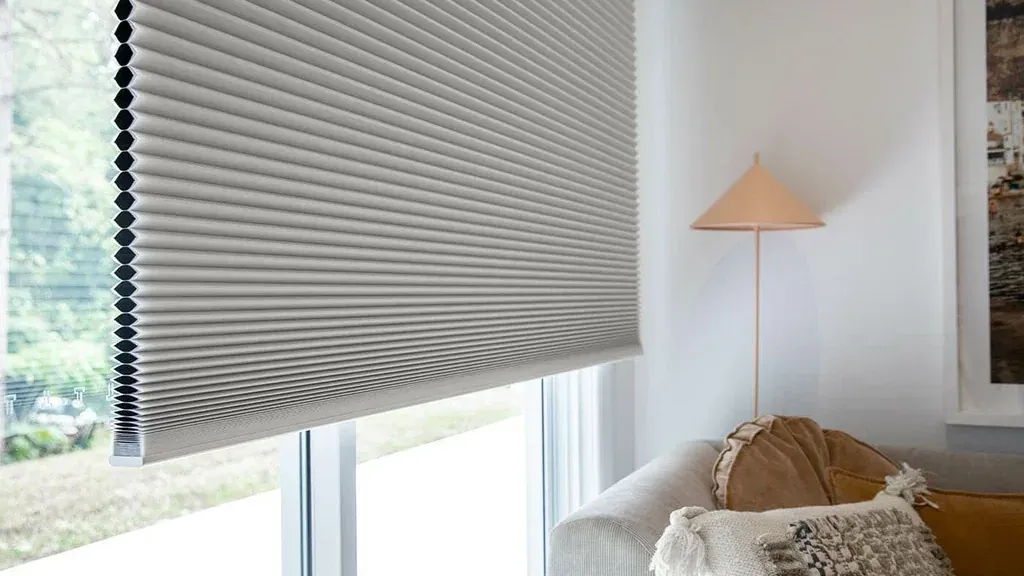A grey honeycomb blind lowered most of the way down a tall living-room window, its pleated cells lit from the side