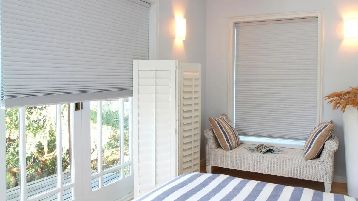 White honeycomb blinds covering a bedroom window and the glazed door beside it, with a folded plantation shutter panel standing between the two