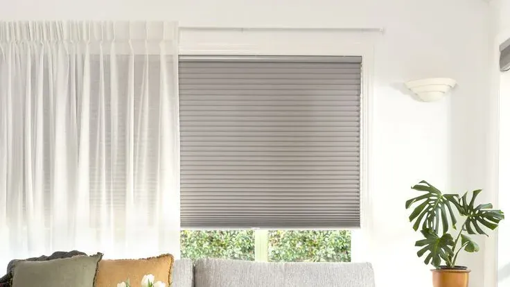 A grey honeycomb blind covering a living-room window, with a white sheer curtain drawn to one side of it and a monstera beside the sofa