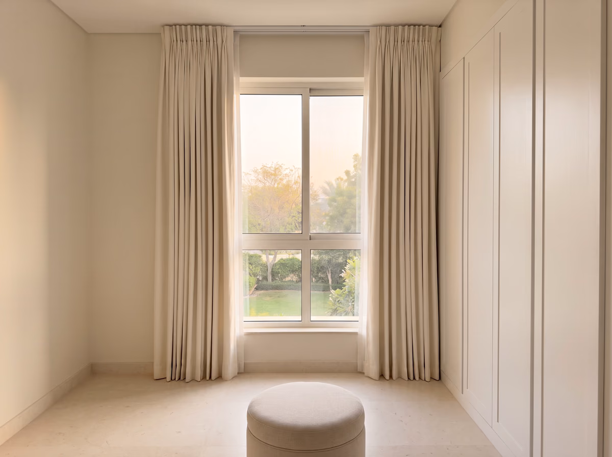 Ivory pinch-pleat curtains drawn back from a villa dressing room window onto a garden, beside fitted wardrobes and a round stool