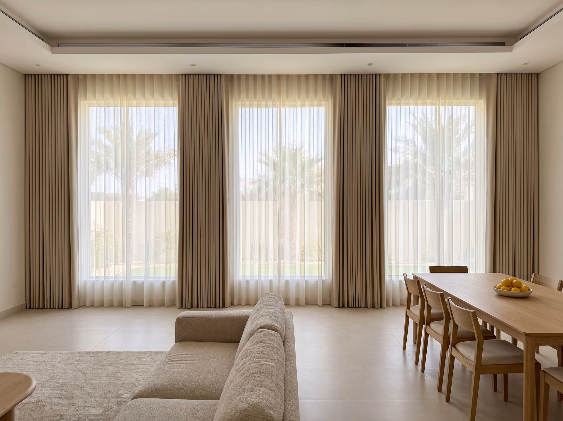 Open-plan villa living and dining room with ivory sheers drawn across three tall windows onto palms and sand curtains stacked between them