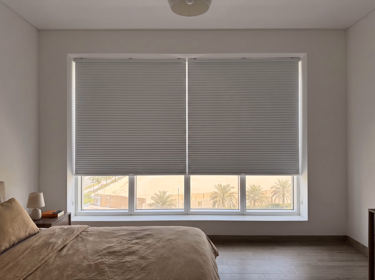 Two light grey blackout honeycomb blinds lowered inside a bedroom window recess, daylight at their edges and palms below them