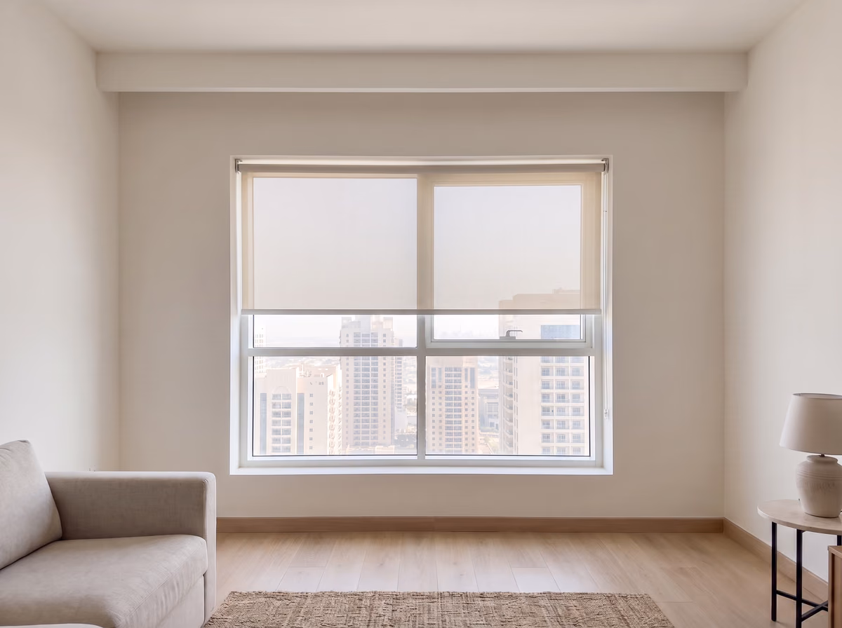 Light grey sunscreen roller blind lowered halfway inside a living room window recess, the towers showing faintly through the fabric