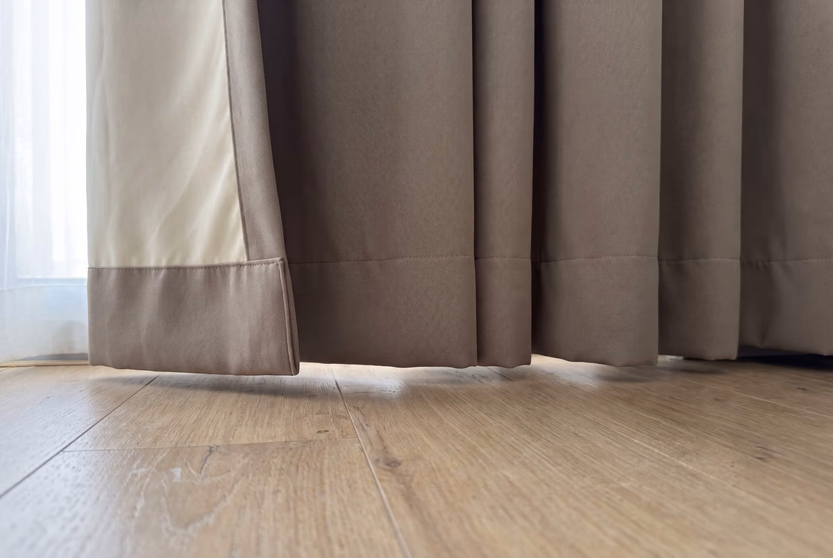Hem of a taupe curtain hanging just clear of a wooden floor, its cream lining showing where the leading edge turns