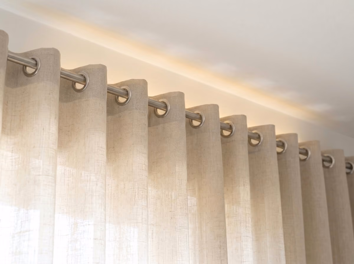 Off-white linen curtain with an eyelet heading on a brushed-steel pole, daylight glowing on the wall and ceiling above the pole