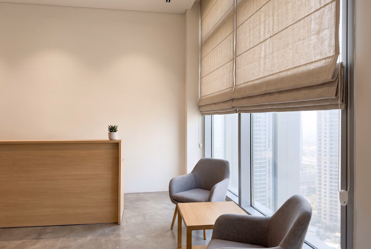 Natural linen roman blinds half raised over the windows of an office reception with an oak desk and two armchairs