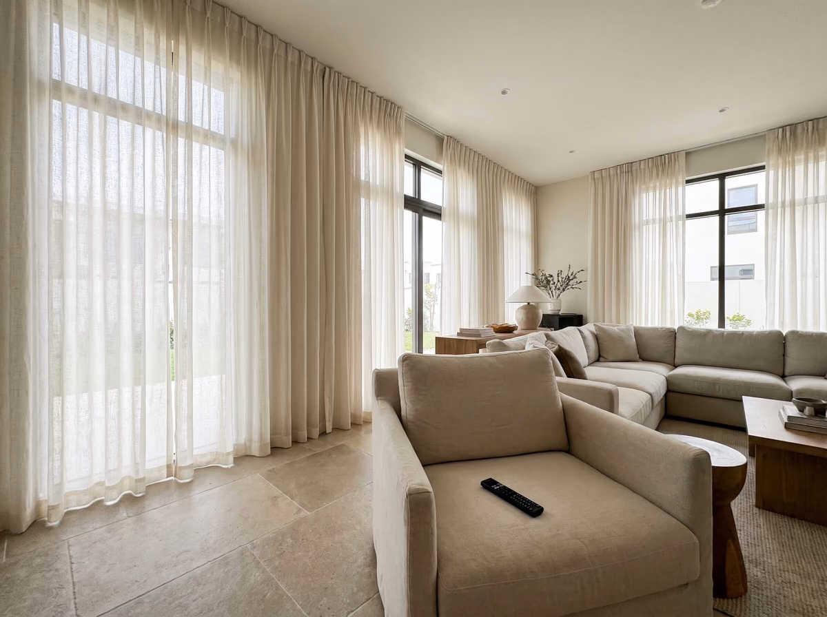 Villa living room with linen-look sheer curtains in a pinch pleat heading at three tall windows, a beige sofa in front and a garden wall outside