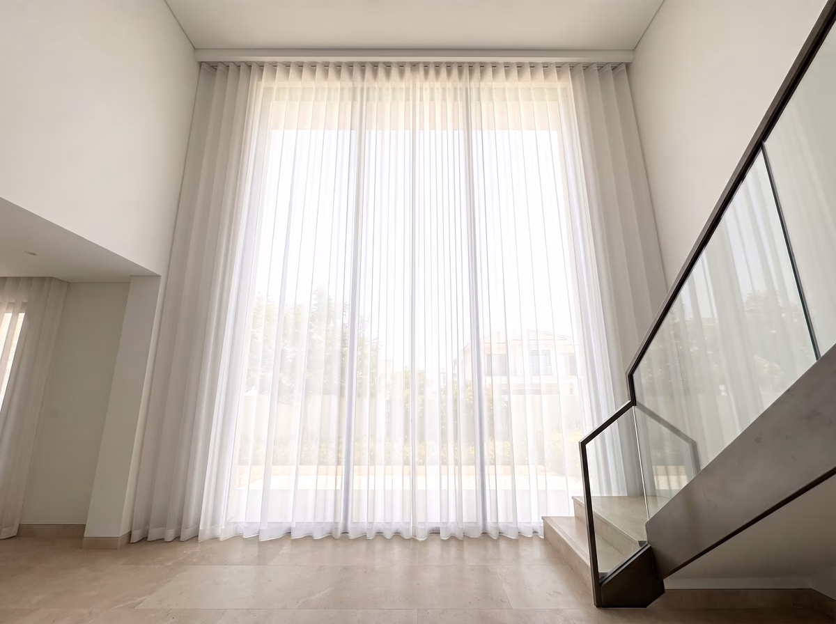 White sheer curtains hanging the full height of a double-height villa window beside a staircase with a glass balustrade