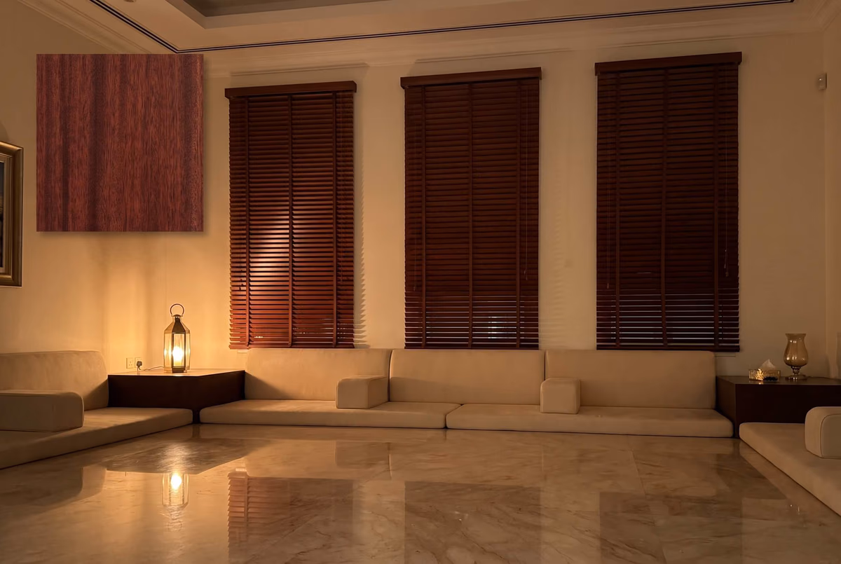 Majlis at night with mahogany wooden blinds closed on three tall windows above floor seating, a lantern lit on a side table and a swatch of the finish inset at top left