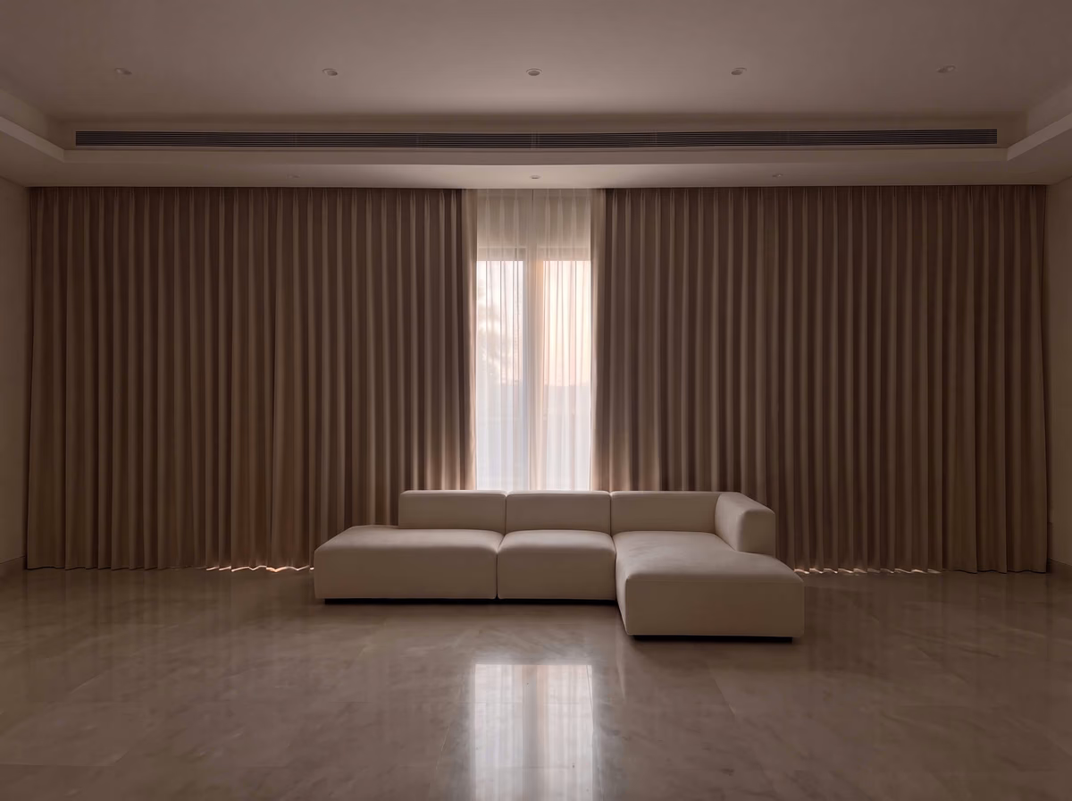 Villa living room at dusk with floor-to-ceiling curtains closed along a long glass wall, a sheer at the centre and a cream sofa on a marble floor