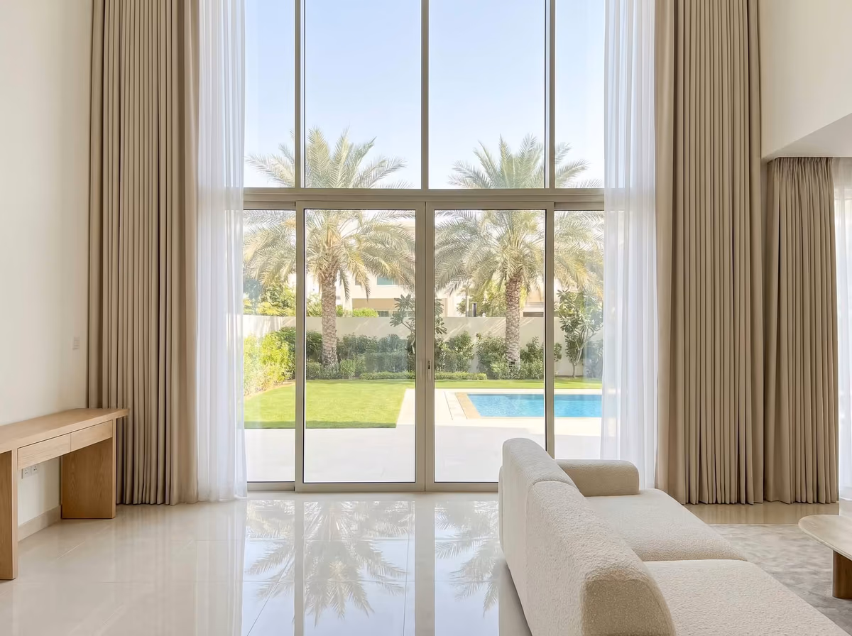 Double-height villa living room with floor-to-ceiling glass, tall beige ripple fold curtains and white sheers drawn back to a garden with date palms and a pool