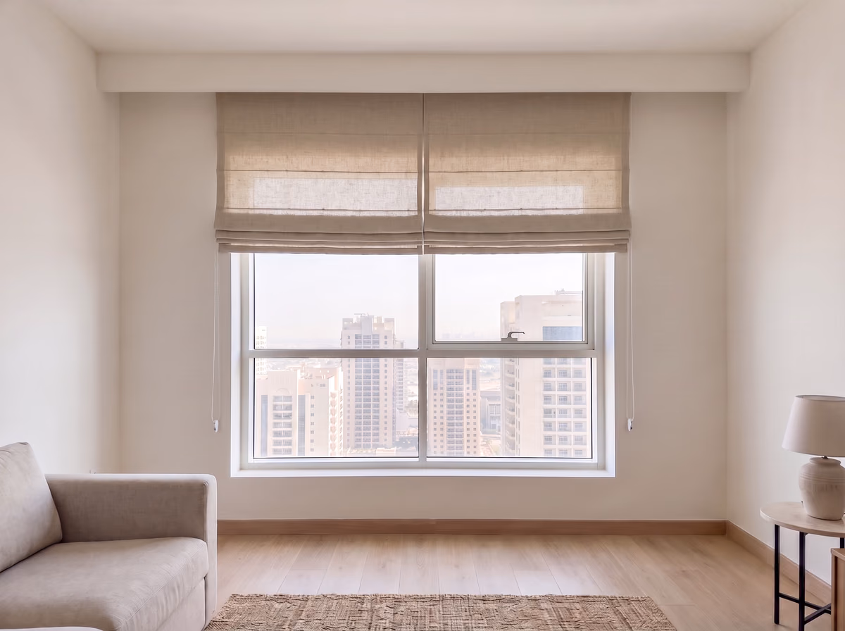Two natural linen roman blinds raised inside a living room window recess, their folds stacked at the top above the towers