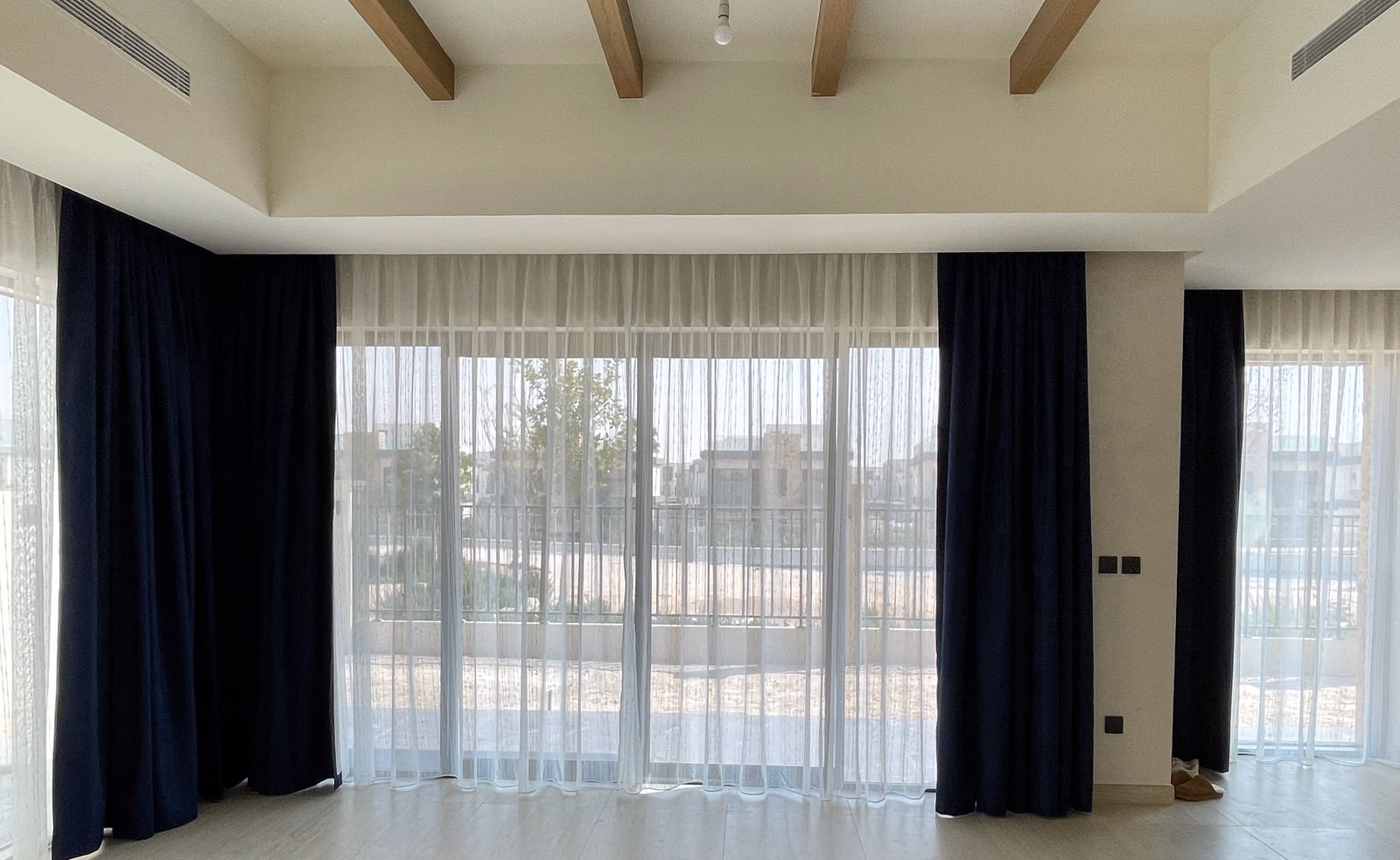 Navy blackout curtains stacked beside a villa's sliding garden doors, white sheers drawn across the glass under a beamed ceiling