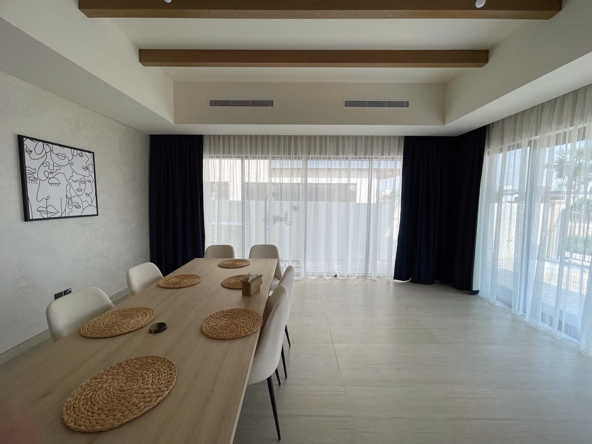 A villa dining area with a long wooden table, navy blackout curtains stacked at the corners and white sheers drawn across glass on two walls