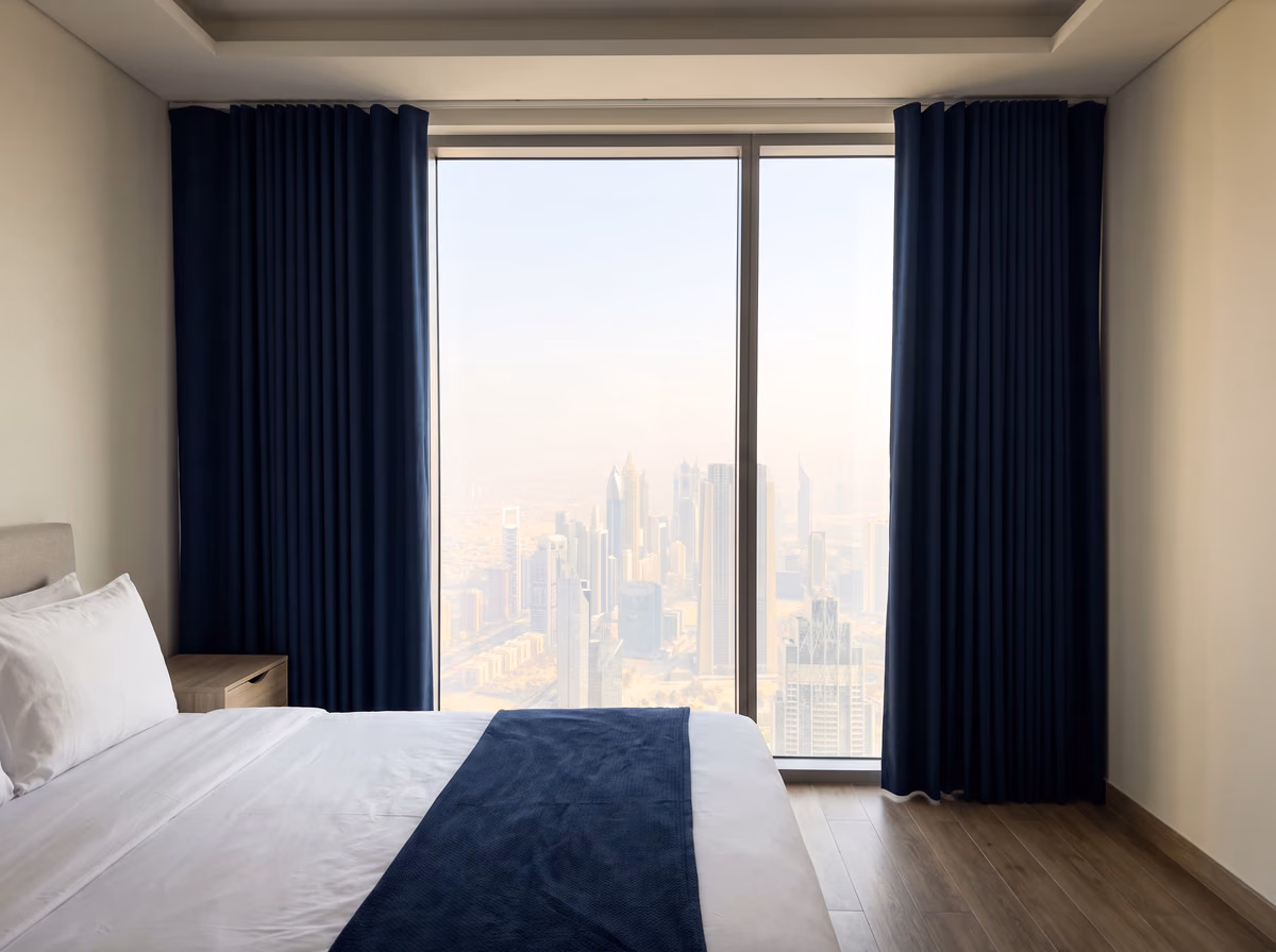 Navy curtains drawn back from a floor-to-ceiling tower bedroom window, a city of towers in the haze beyond a made bed