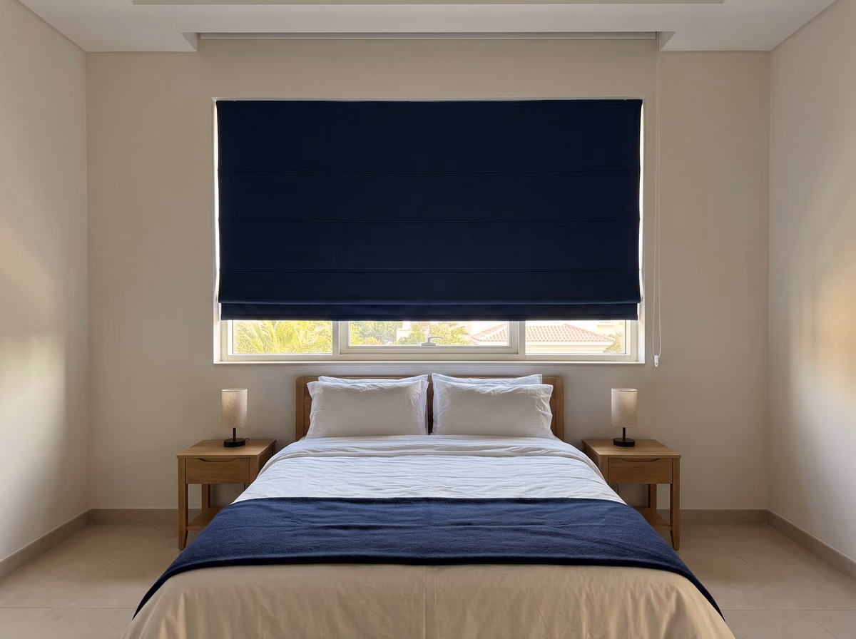 Navy blackout roman blind lowered over most of a bedroom window above a made double bed, palms and a tiled roof below it