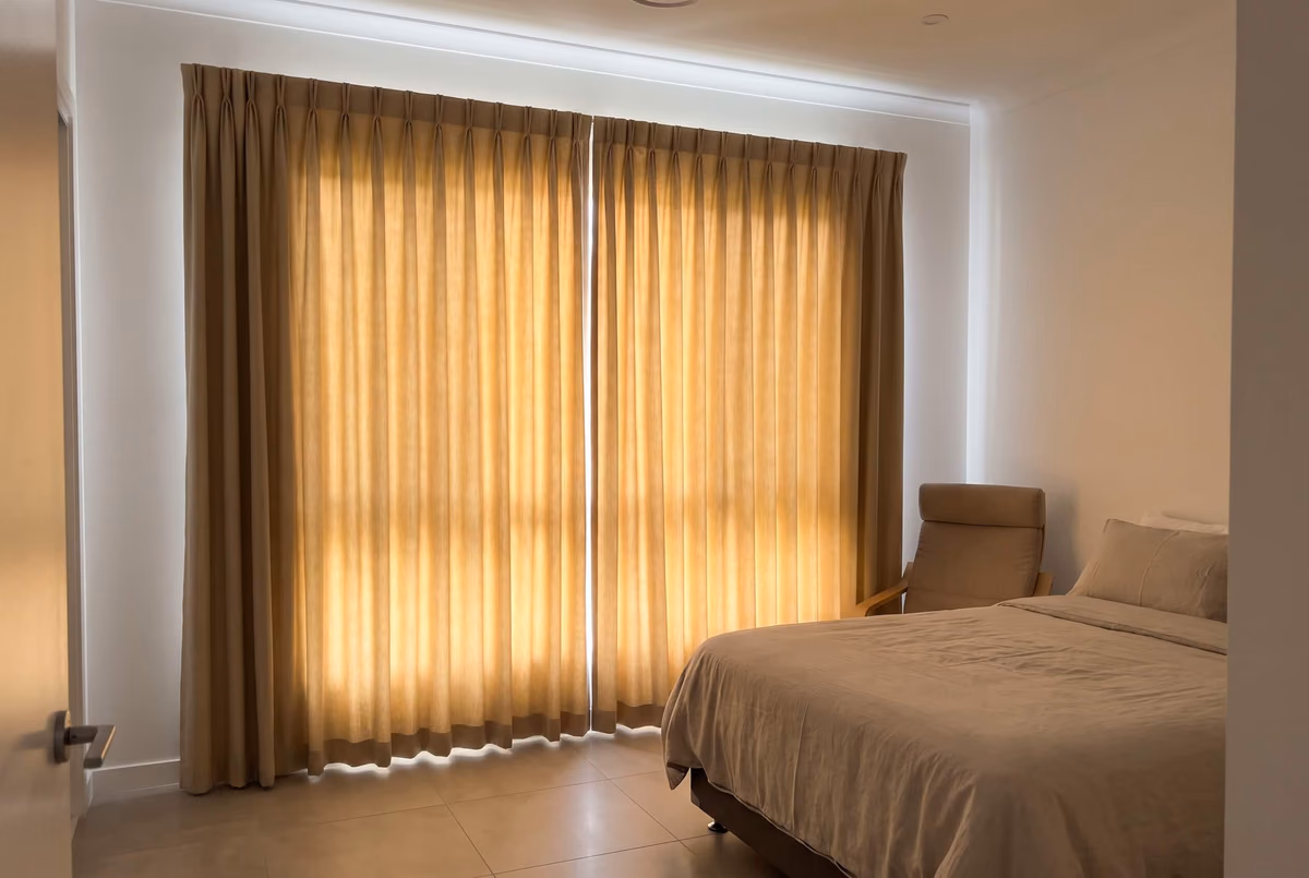 Ochre pinch-pleat curtains closed across a bedroom window, low sun glowing through the cloth
