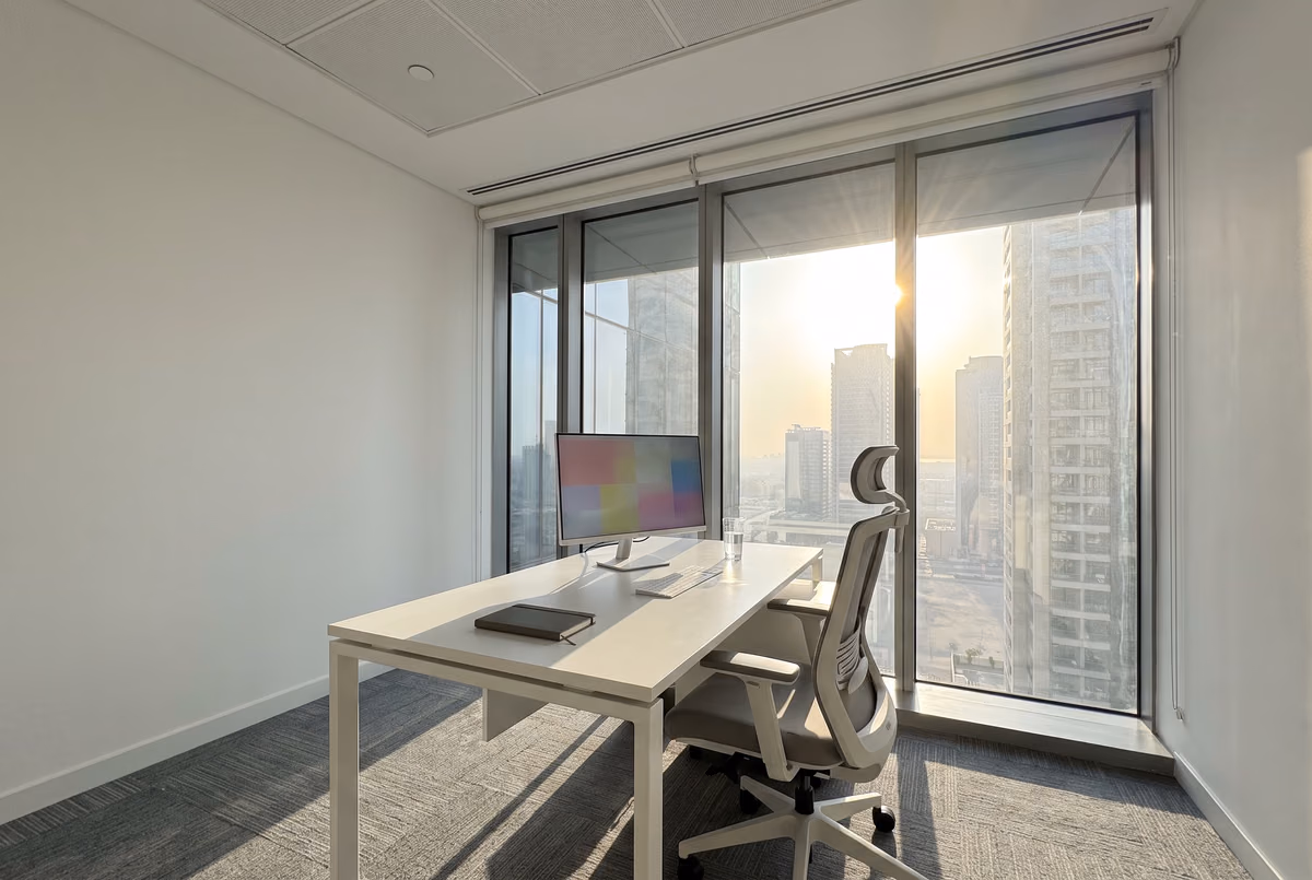 Office desk beside floor-to-ceiling glass in the afternoon, the blinds raised and low sun pouring across the desk and the screen