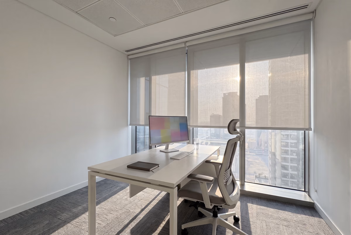 The same desk with sunscreen roller blinds lowered over the glass, the sun softened and the towers still visible through the fabric