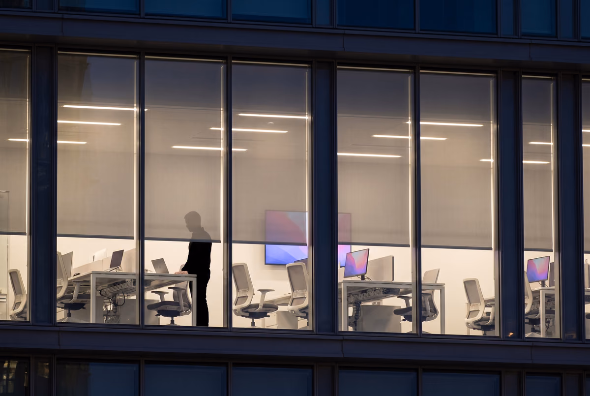 The same office floor at night with the lights on, desks, monitors and a standing figure visible through and below the lowered blinds
