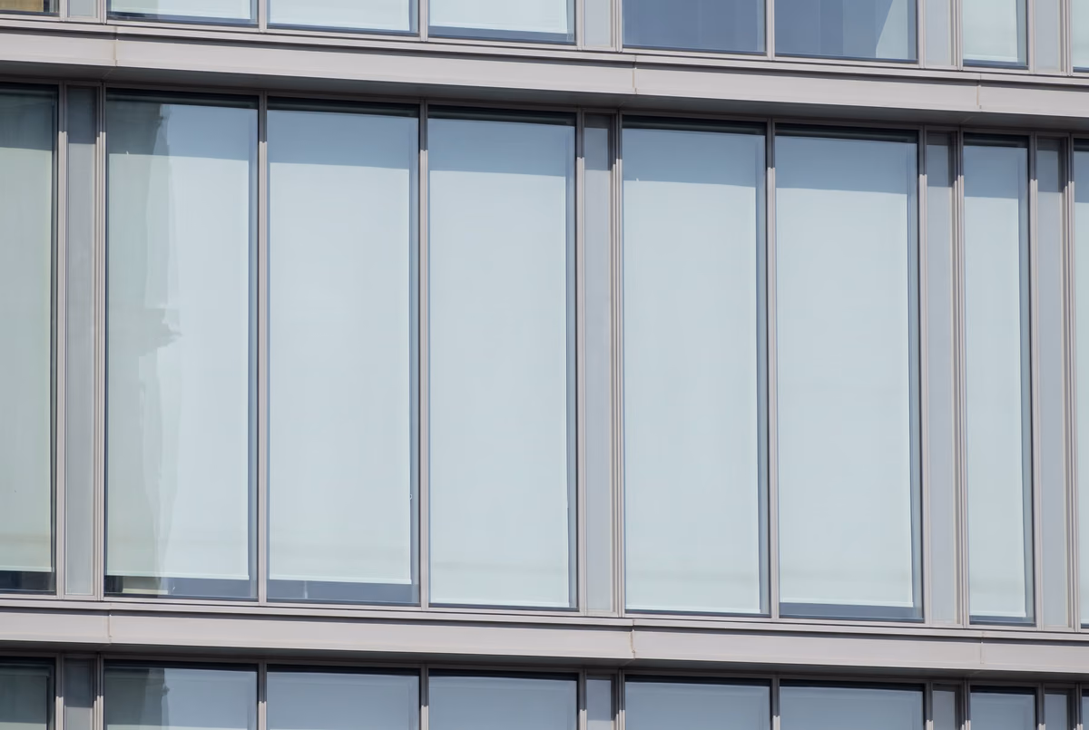 One floor of a glass office tower seen from the building opposite at midday, sunscreen blinds lowered and the windows reading as pale panels