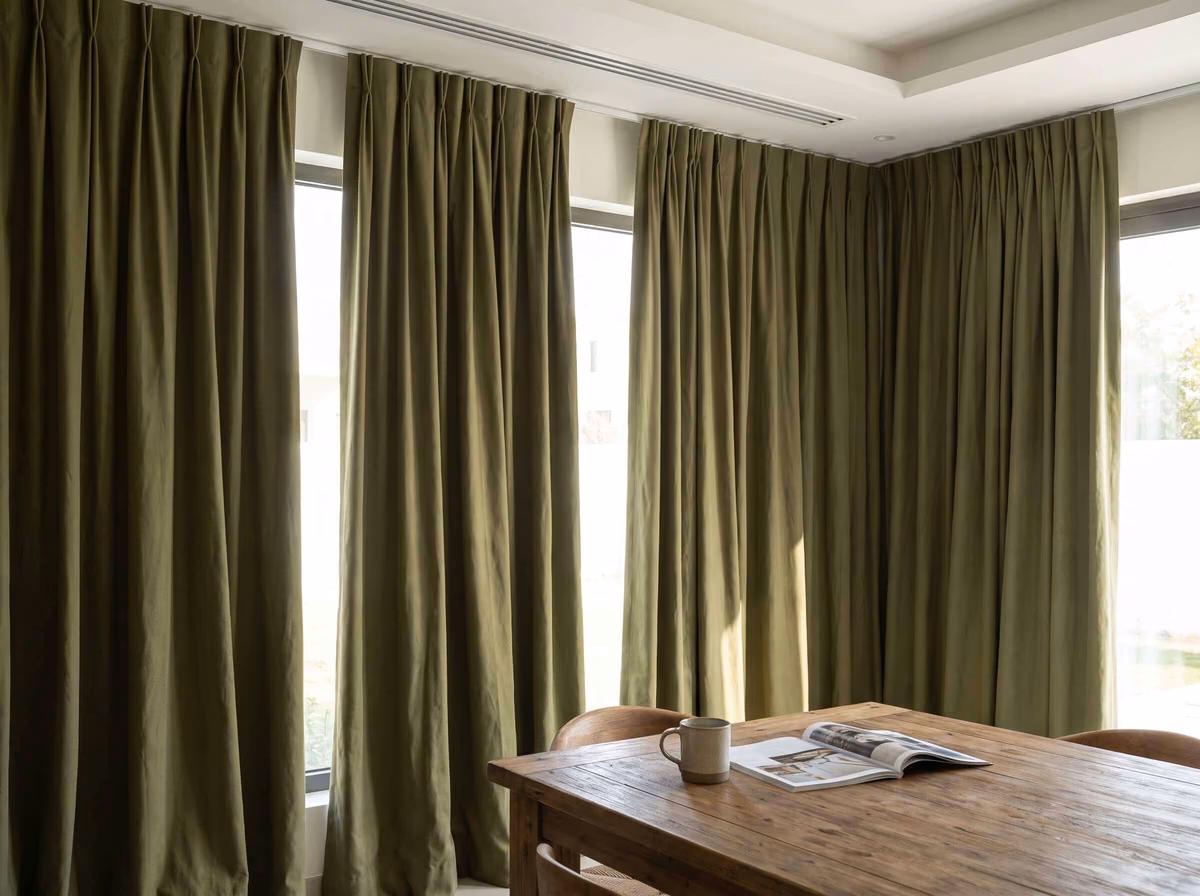 Olive-green pinch-pleat curtains closed across the corner windows of a villa dining room, a wooden table in front