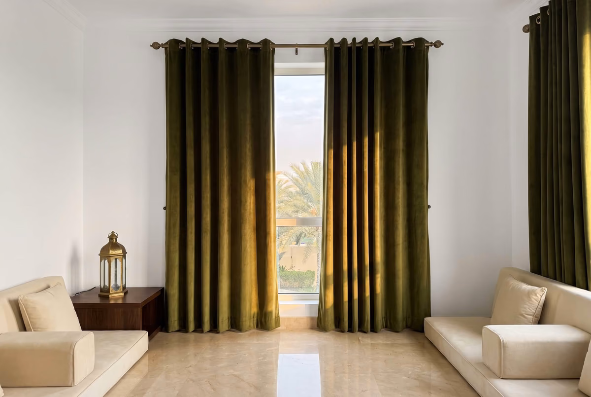Olive velvet eyelet curtains on an antique brass pole with round finials, beside a floor-seating majlis and a brass lantern