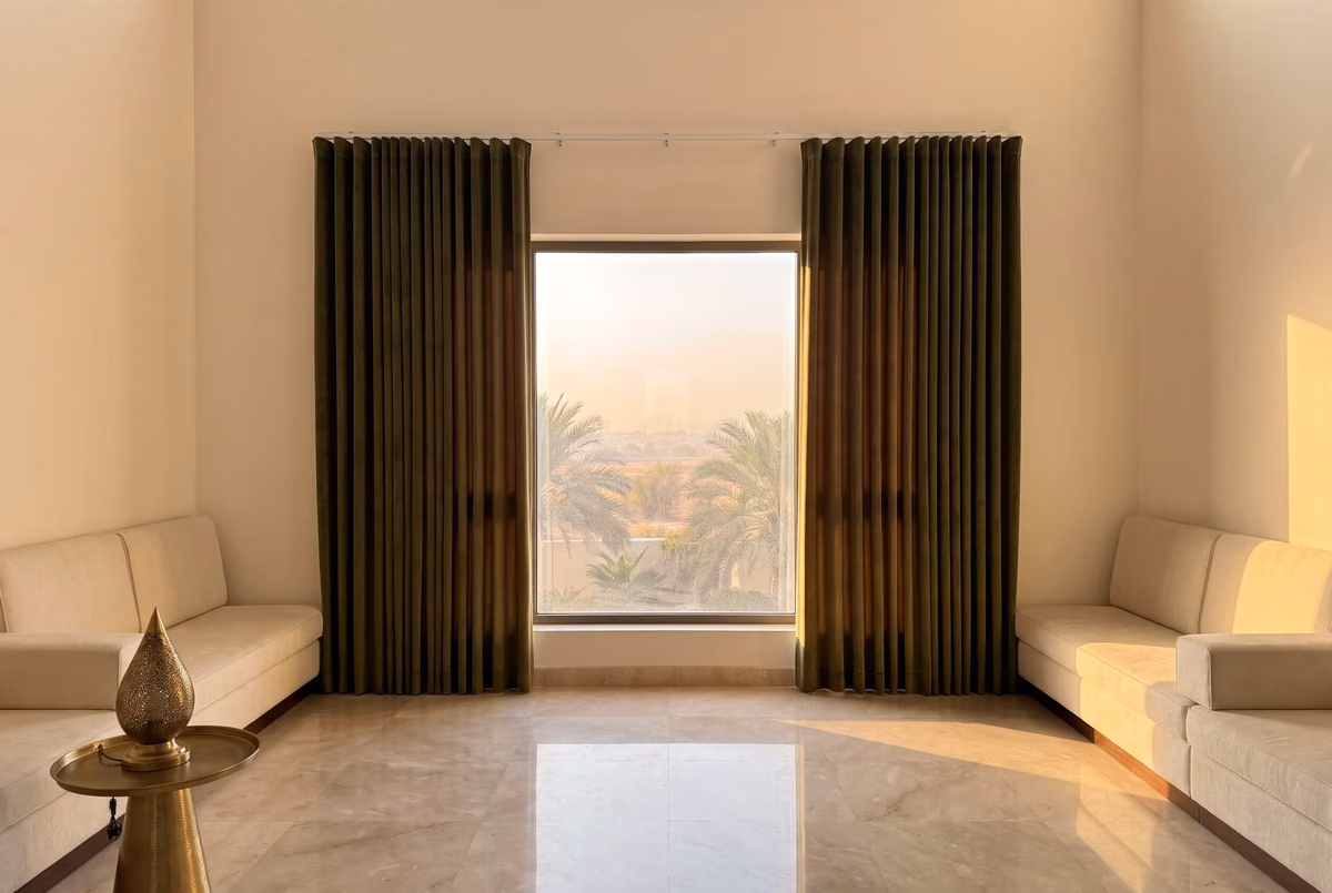 Majlis with low cream seating and deep olive wave curtains drawn to each side of a tall window, palms outside, a brass lantern on a table