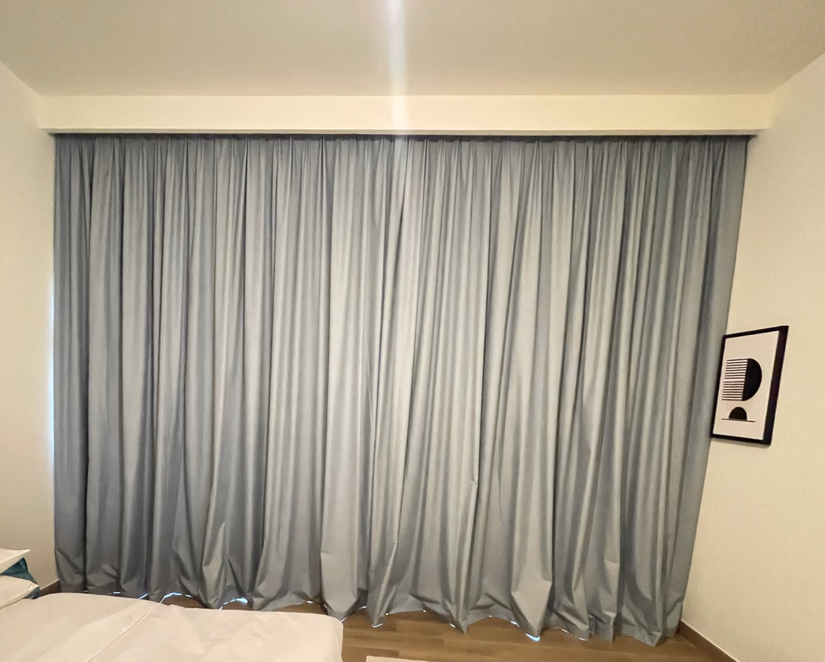 A pale blue-grey blackout curtain drawn wall to wall across a bedroom window, from the ceiling to the floor, beside a white bed