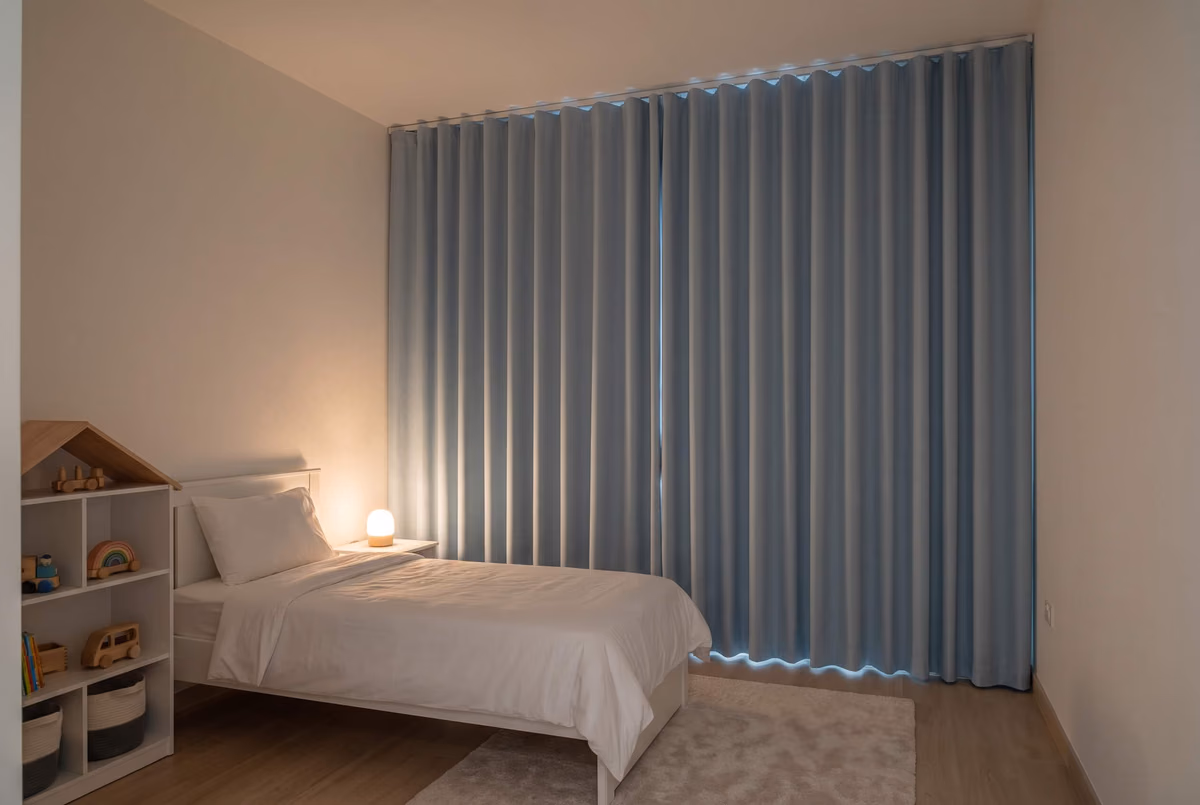 Child's bedroom at night, pale blue blackout wave curtains closed with a little light above and below them, a small lamp lit by the bed