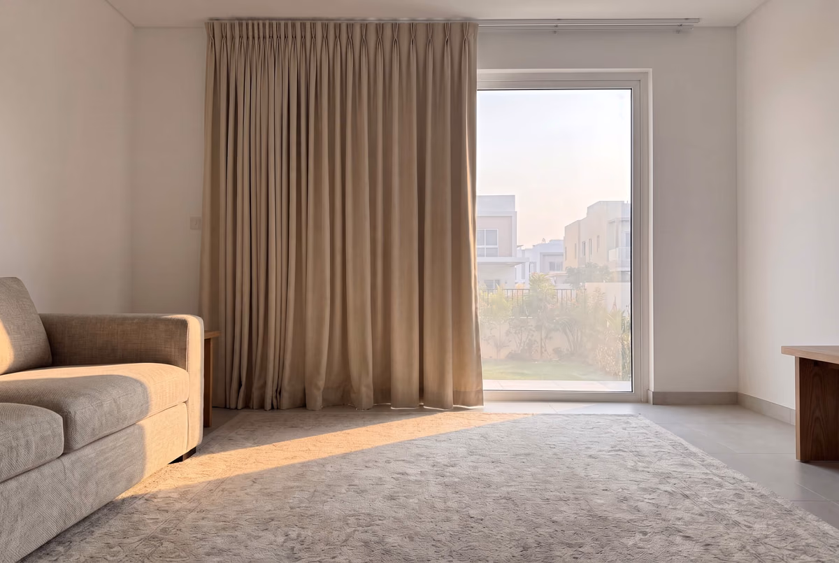Taupe pinch-pleat curtain drawn back onto the wall beside a garden door, afternoon sun lying across the rug