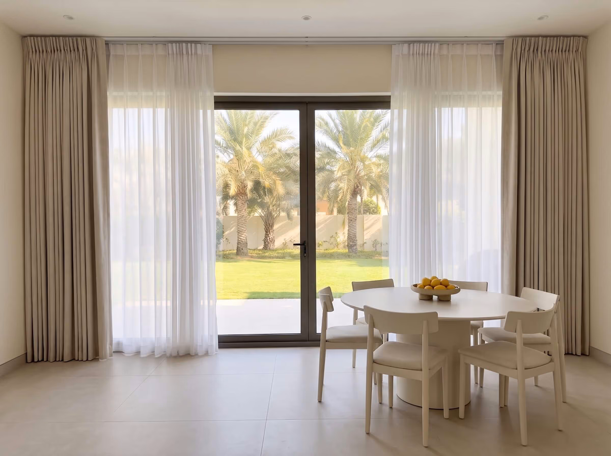 Beige pinch pleat curtains and white sheers on a ceiling track around glass doors to a garden of date palms, a round dining table in front