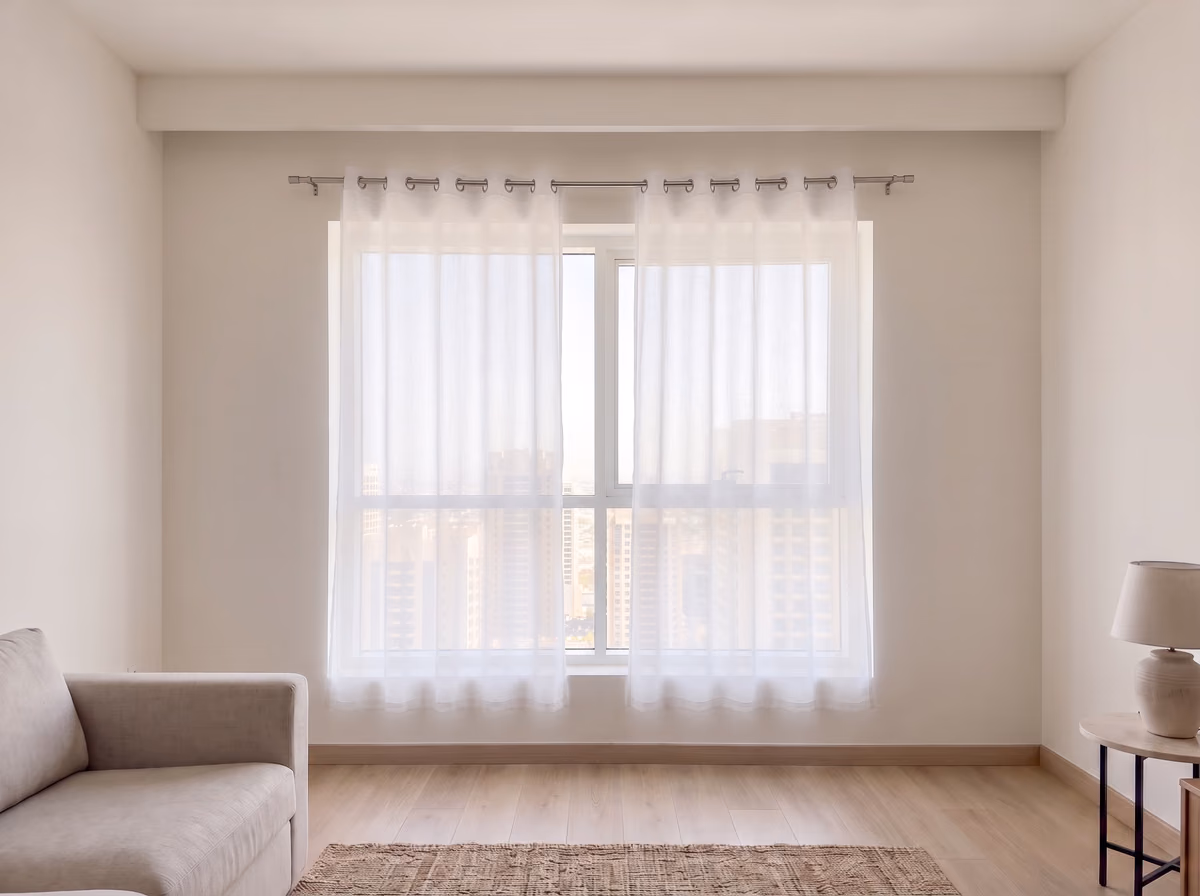 Two ready-made white sheer panels on a thin rod above a living room window, stopping short of the floor and of the window's sides