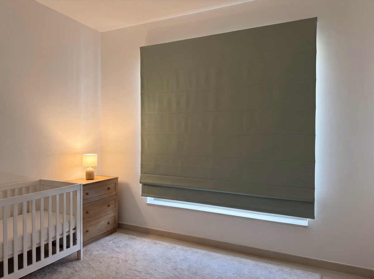 Nursery with a large sage green roman blind lowered over the window, a white cot beside it and a small lamp lit on a wooden chest