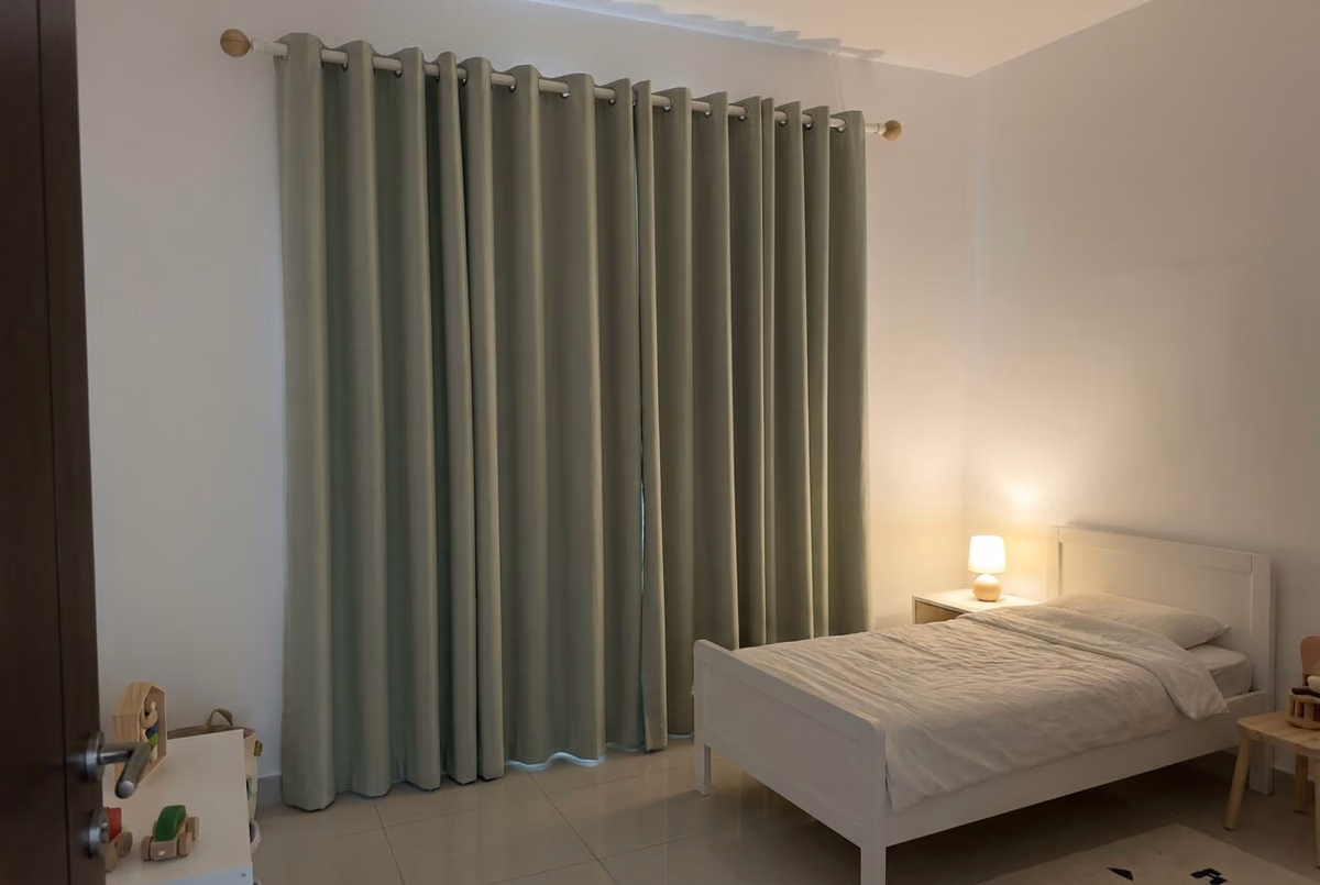 Child's bedroom at night with sage green eyelet curtains closed on a white pole, a small lamp lit beside the bed