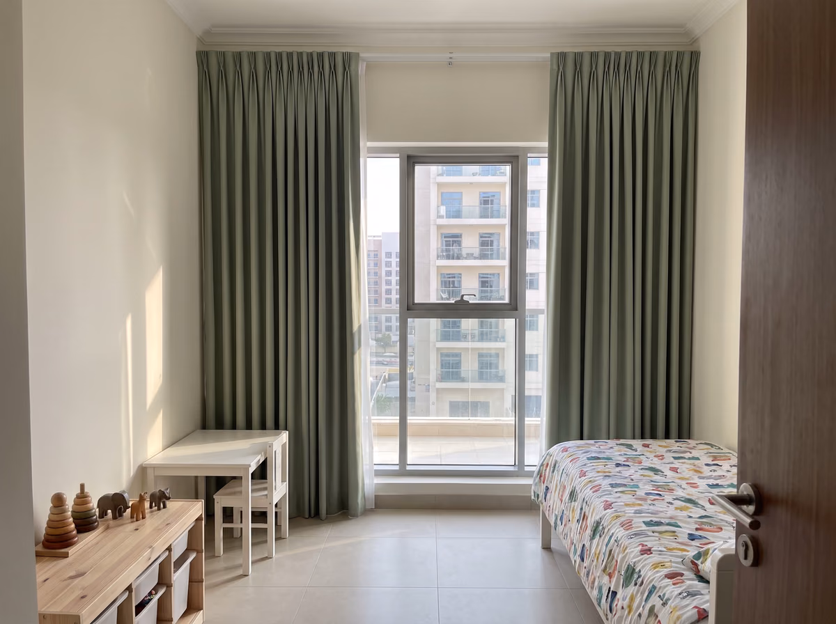 Children's bedroom with sage green pinch-pleat curtains drawn back from a tall window above a small white desk and a single bed