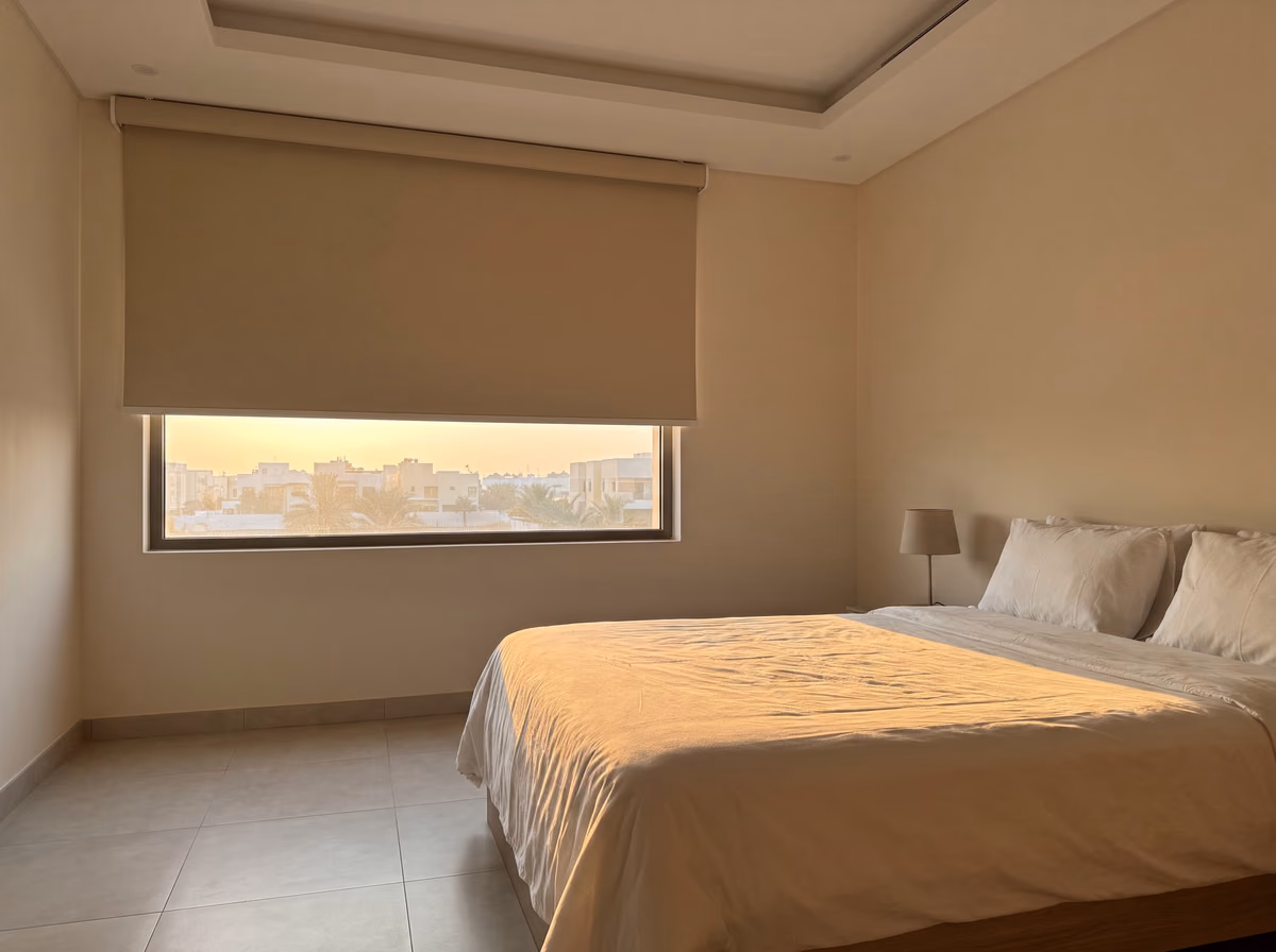 Villa bedroom at sunrise with a sand-coloured blackout roller blind raised part way, warm low light falling across the bed