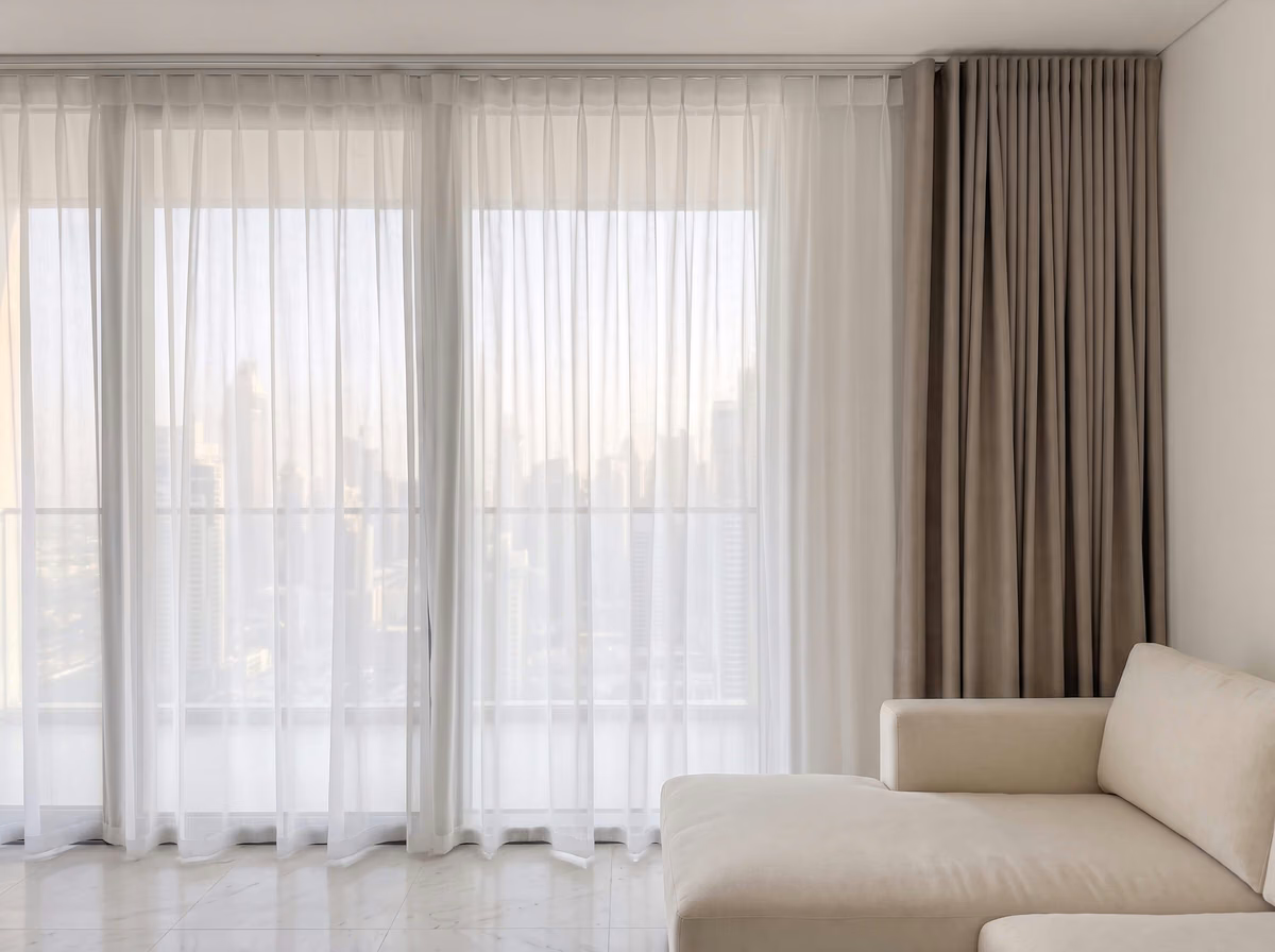High-rise living room with a white sheer drawn across the glass and a taupe ripple fold curtain stacked at the side, city towers beyond