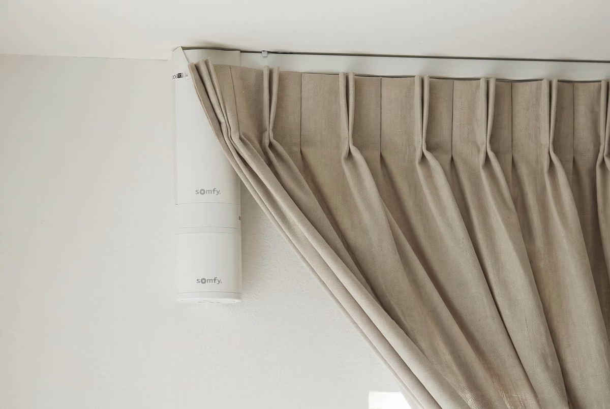 Close-up of a white motor marked Somfy at the end of a ceiling track, a beige pinch pleat curtain hanging beside it