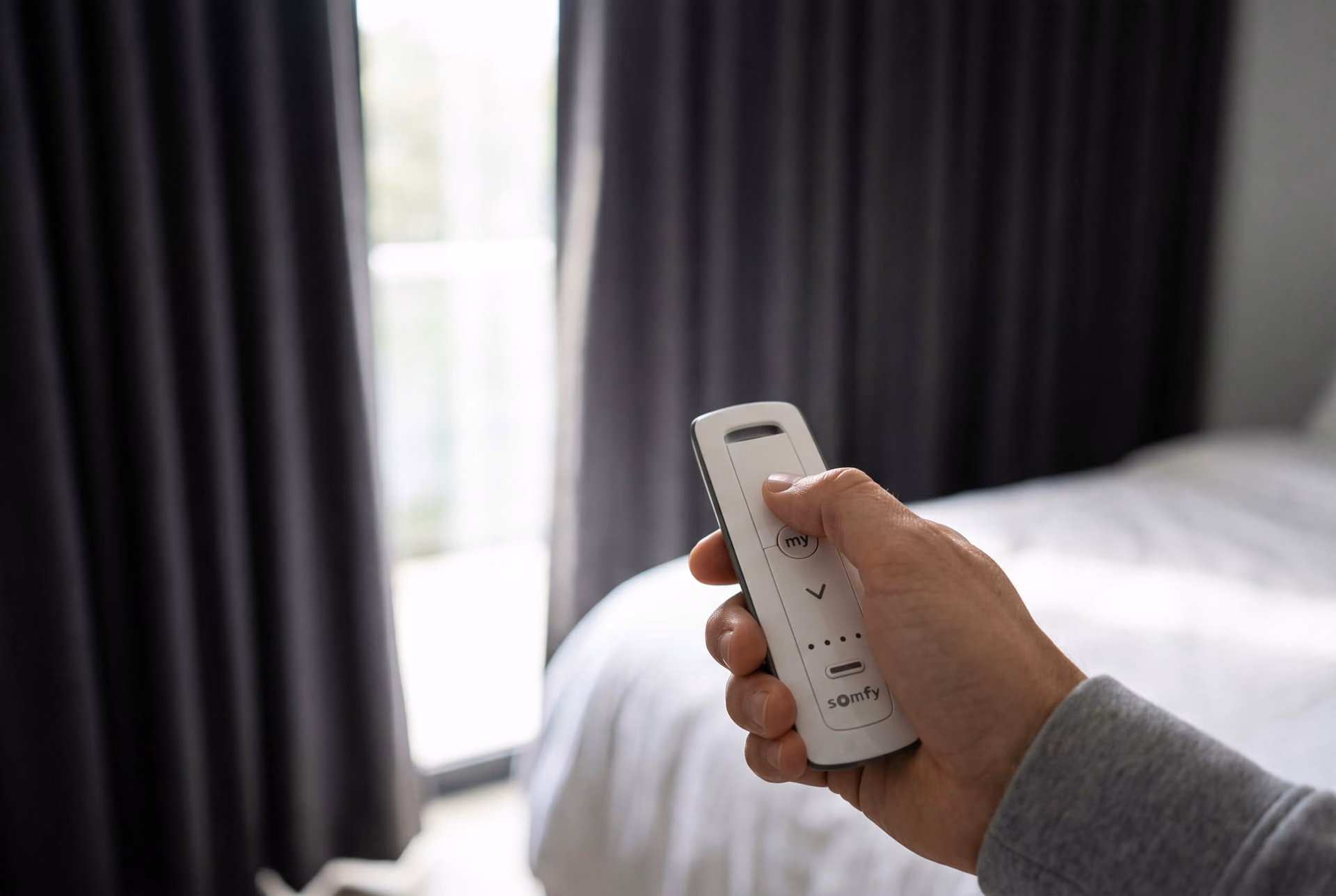 A hand holding a white Somfy remote beside charcoal blackout curtains drawn across a bright window, a white bed alongside