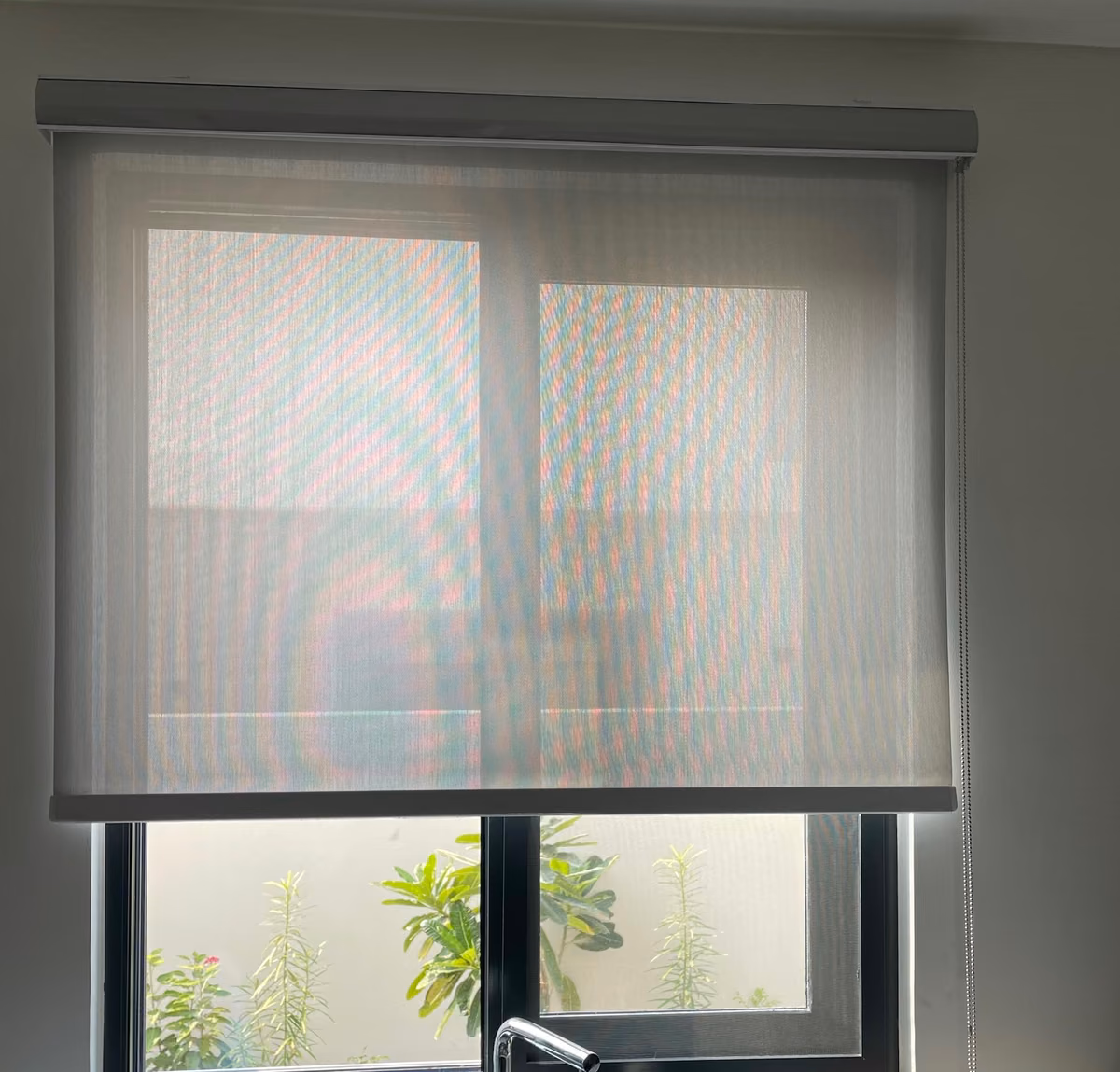 A grey sunscreen roller blind pulled halfway down a kitchen window above the sink, green planting visible through the uncovered lower glass