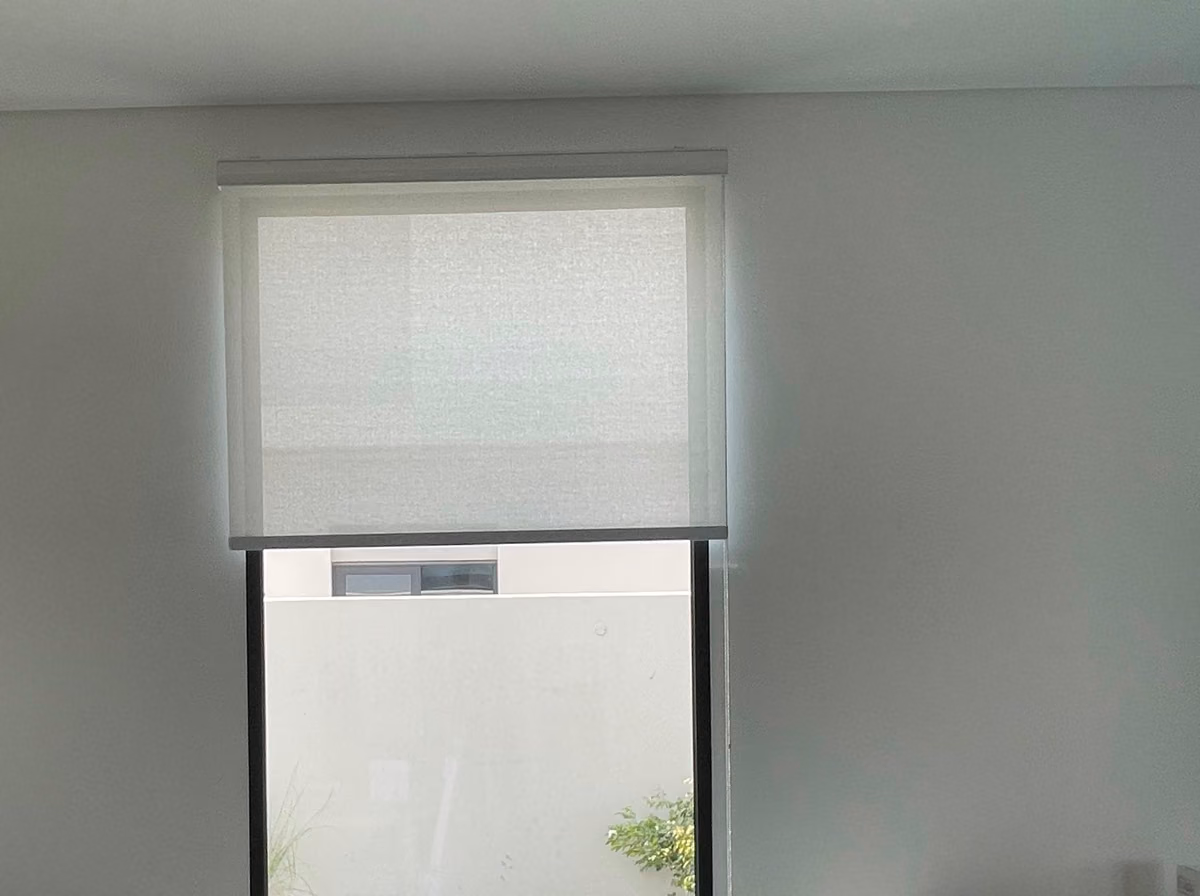 A single sunscreen roller blind lowered halfway over a tall glass door in a villa dining area, a walled garden and lawn beyond it