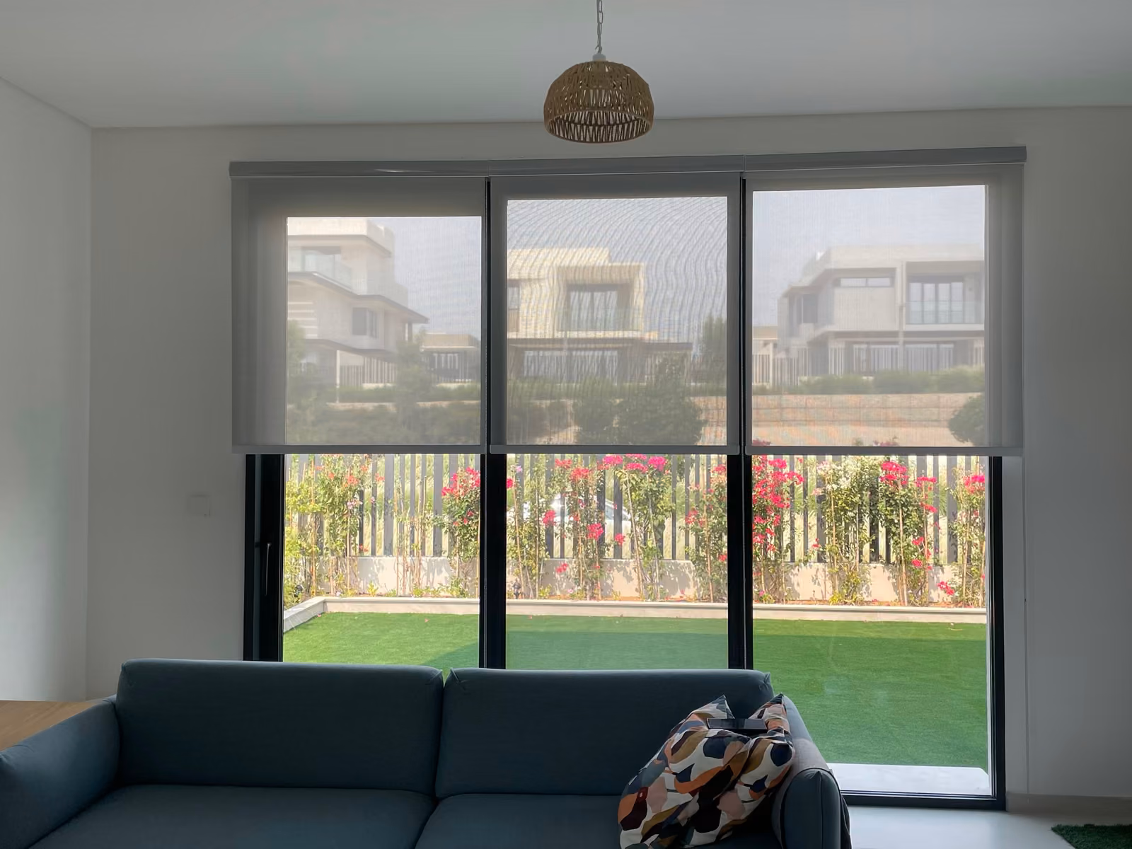 Three grey sunscreen roller blinds half lowered over a villa's triple glass doors, the villas opposite showing through above a garden of bougainvillea