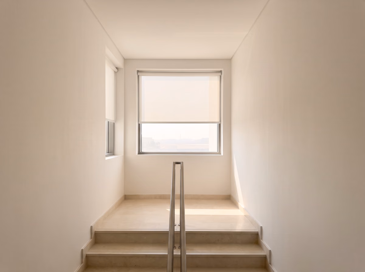 Stairwell landing with a white sunscreen roller blind half lowered inside the window recess, a second roller on a narrow side window