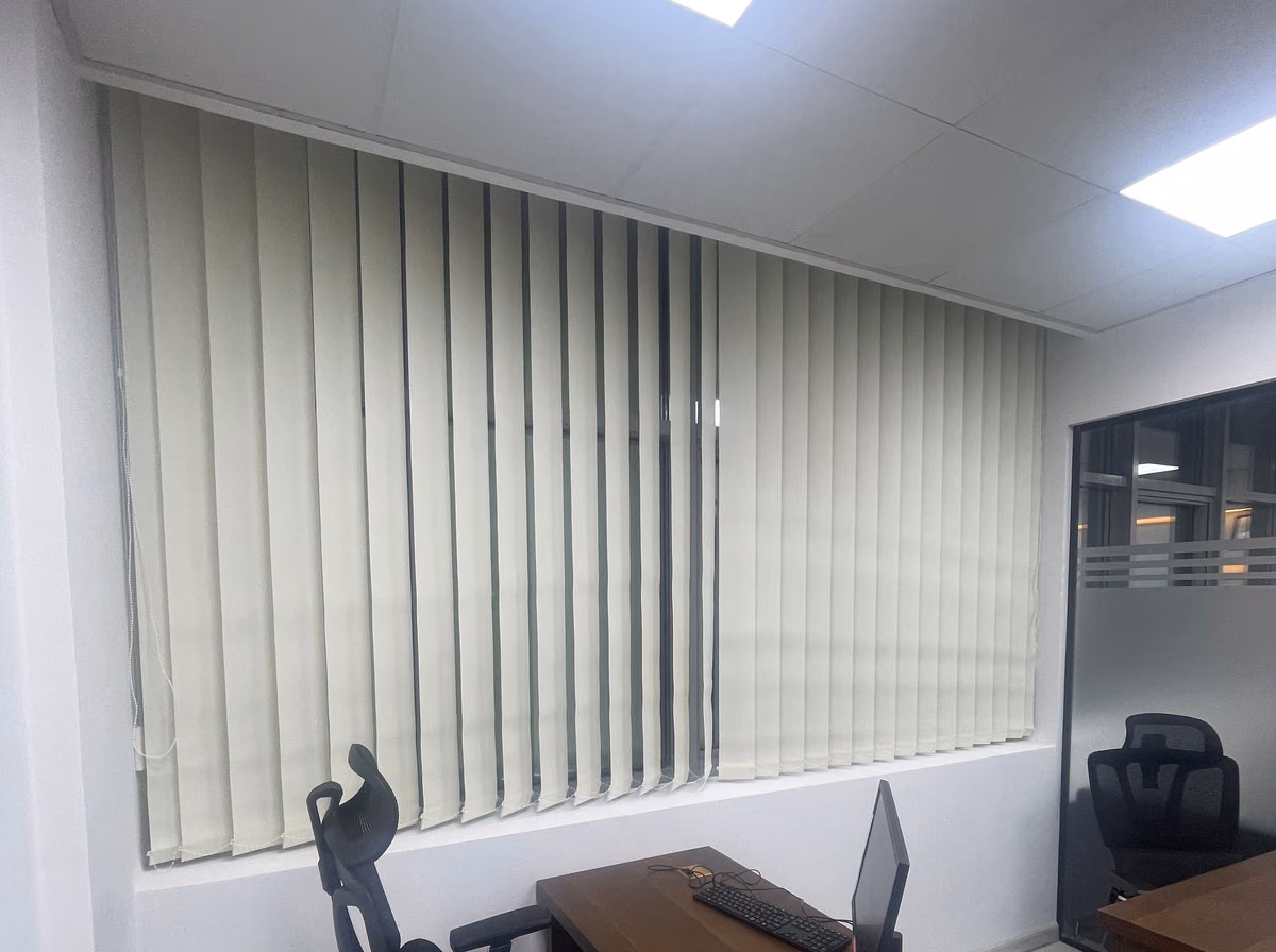 Sunscreen vertical blind louvres turned part open across a dark office window at night, a desk and office chair below