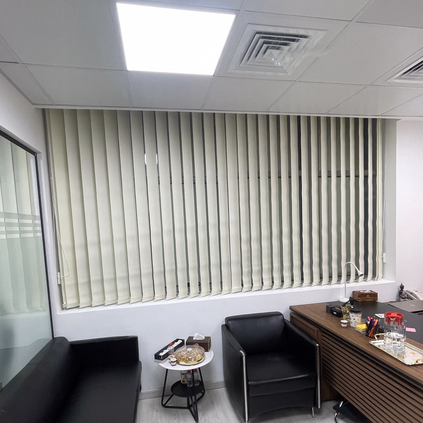 Off-white sunscreen vertical blinds across a manager's office window above a black leather sofa and armchair, a ceiling air-conditioning vent close to the louvres