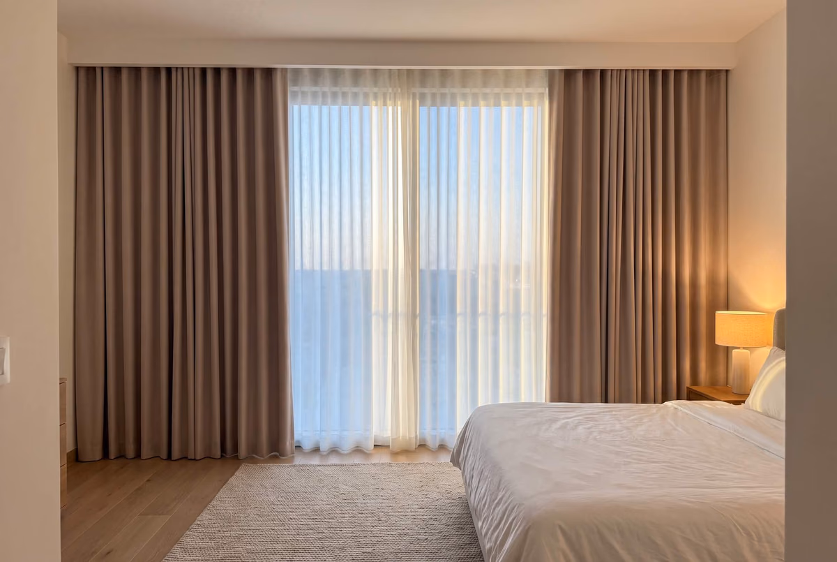 Bedroom with taupe blackout wave curtains drawn back and a white sheer wave closed across the glass between them, a bedside lamp lit